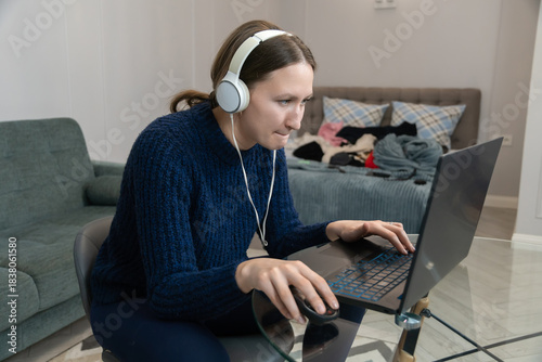 Focused young woman wearing headphones works from home, typing on her laptop and using a mouse, while a cluttered bedroom creates a chaotic backdrop to her everyday life