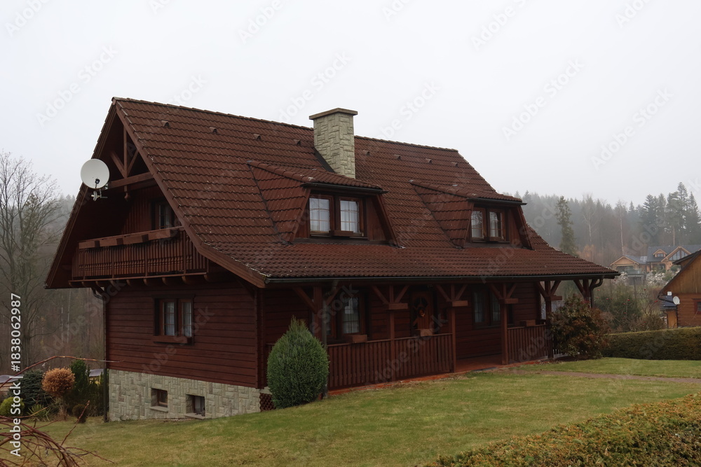 Obraz premium Traditional Wooden Cottage with Red Tiled Roof and Stone Chimney, Rustic Mountain House Exterior with Balcony and Porch, Rural Architecture in Foggy Weather, Classic Timber Home Facade