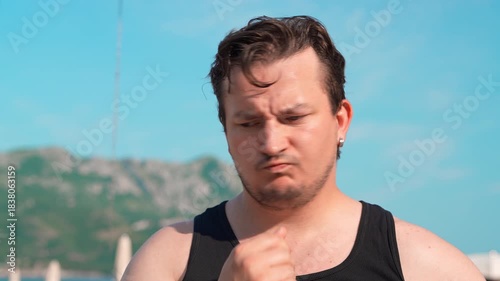 A man in a black T-shirt stands outside under a clear blue sky, his hand to his chin, a pensive expression on his face. Hills are barely visible in the background.