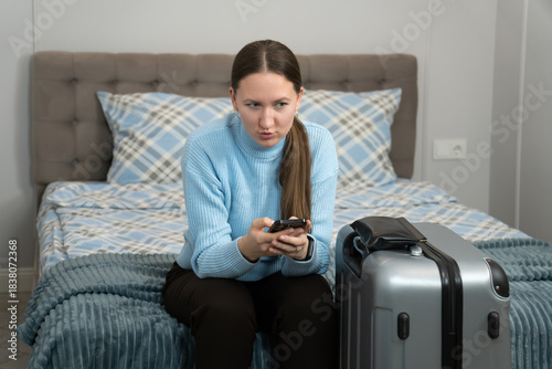 Businesswoman sitting on a bed next to a packed suitcase, using a smartphone to finalize plans for an upcoming business trip or vacation, blending work and travel seamlessly