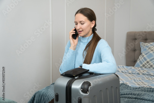 Young woman is making a phone call while sitting on a bed with her packed suitcase, likely confirming travel details or saying goodbye before leaving for a trip