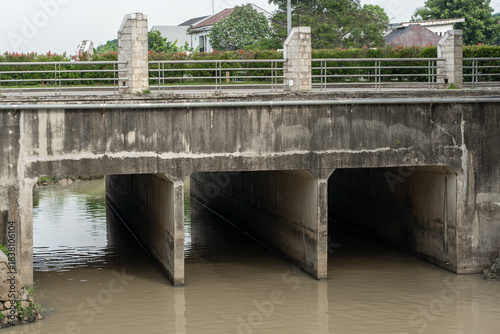 Concrete bridge with box culverts and metal railings crossing a murky river for urban flood control and drainage infrastructure.
