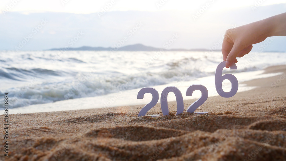 Naklejka premium hand place the number 6 on sandy beach, transition from year 2025 to 2026 for new year sign on summer beach at sunset time, with ripple sea wave and beautiful nature at background, New Year celebrate