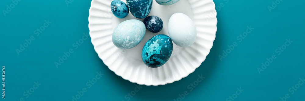 Fototapeta premium Colorful blue Easter eggs are artistically arranged on a white plate resting on a vibrant teal surface, capturing the essence of spring festivities and holiday traditions, banner