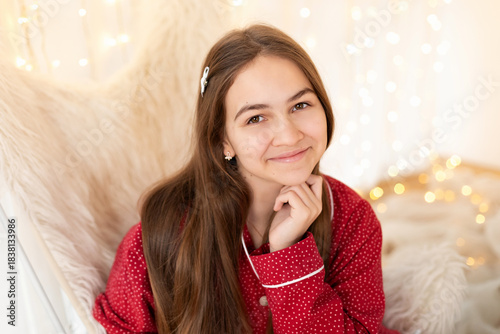 Closeup portrait young cute girl in red festive pajamas sit on armchair near  glowing lights garlands at home. Portrait Happy smiling teen on Xmas Eve. New year mood. 
