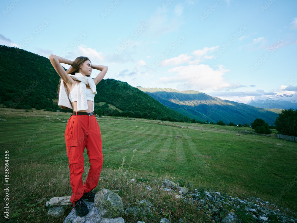 Naklejka premium Outdoor fashion model in a mountain field wearing red pants and white top, hands raised, carefree pose under blue sky, showcasing nature inspired style and rural landscape