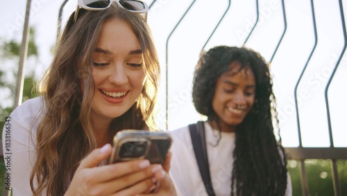 three multiracial female friends one disabled in a wheelchair enjoy a sunny day outdoors while laughing and sharing moments on their smartphones at a park setting during afternoon