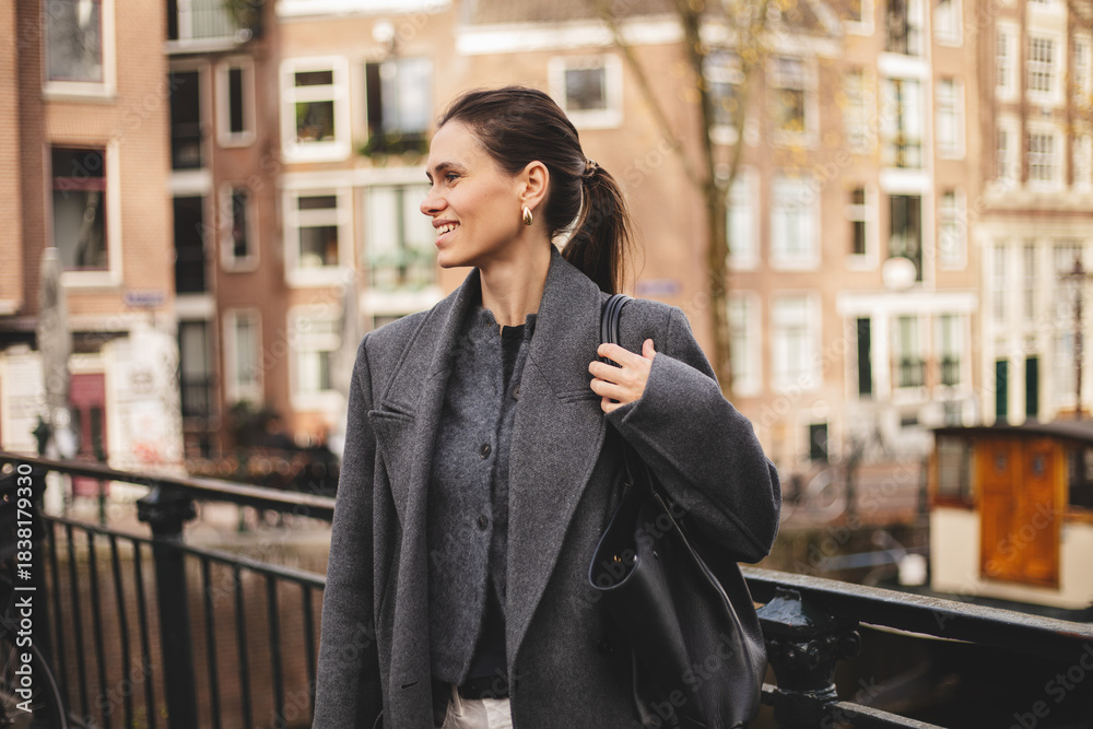 Fototapeta premium Smiling woman walking outdoors in the city. Modern urban lifestyle, casual fashion, natural beauty and confident female portrait. Girl with high ponytail wear grey wool coat and hold black handbag.