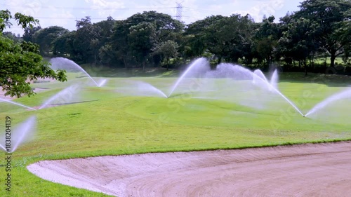 Automatic sprinkler system spraying water across a lush green fairway of a golf course under a clear sky. A sand bunker is visible in the foreground.