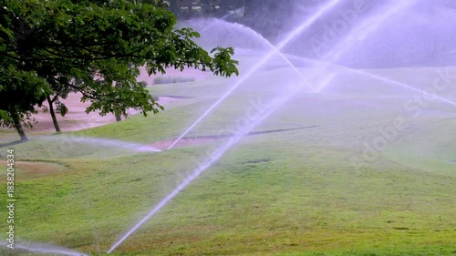 Sprinklers actively irrigating the bright green grass on a golf course, with a curved sand trap occupying the bottom frame. Trees line the background horizon.