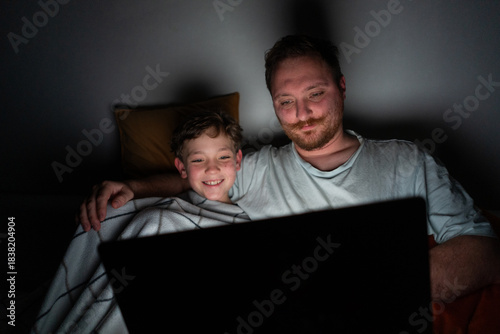 A father and son sit closely on a bed, looking at a laptop screen. The room is dark, with soft light from the laptop illuminating their faces. They are sharing a moment of laughter and joy.
