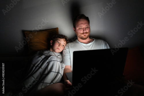 A father and son sit closely on a bed, looking at a laptop screen. The room is dark, with soft light from the laptop illuminating their faces. They are sharing a moment of laughter and joy.