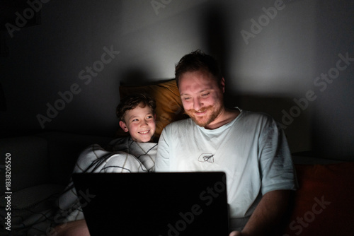 A father and son sit closely on a bed, looking at a laptop screen. The room is dark, with soft light from the laptop illuminating their faces. They are sharing a moment of laughter and joy.