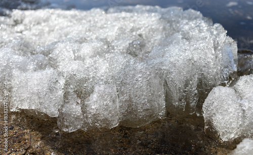 Close-up view of ice formation along a rocky shoreline, showcasing glistening textures and natural beauty, with sunlight reflecting off the ice, creating a serene winter atmosphere