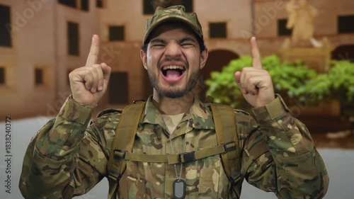 Young hispanic man in camouflage military uniform points finger upward in front of an old town building; pride duty honor loyalty.