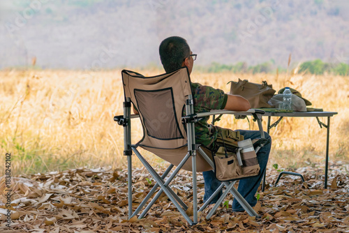 Back view of man tourist sit alone on camping chair in dry field. Man resting after long day of hard farm work, solitude and peaceful contemplation at end of day. Relaxation traveling in park concept.