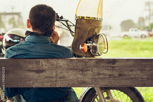 Back view of motorcyclist rests at roadside pavilion and uses smartphone while waiting for rain to stop from heavy rain on side of road. Using communication technology while experiencing travel issues