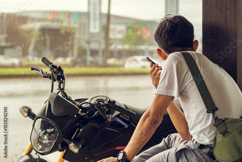 Back view of motorcyclist rests at roadside pavilion and uses smartphone while waiting for rain to stop from heavy rain on side of road. Using communication technology while experiencing travel issues