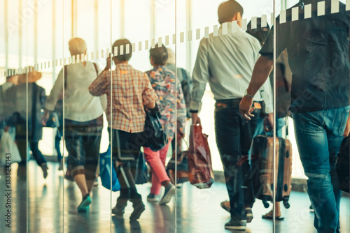 Photograph through large clear glass wall with blurred image of departing passengers and luggage walking airplane boarding corridor from terminal to plane in background. Selective focus on glass.