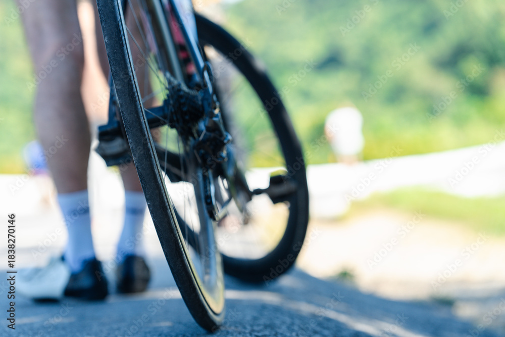 Naklejka premium A close-up view of a bicycle tire with a cyclist's legs in the background, capturing a moment of leisure activity outdoors.