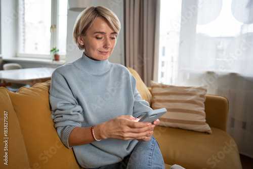 Positive middle aged woman resting on couch in living room with phone, scrolling social networks online, chatting with friends watching videos, enjoying browsing on smartphone while sitting on sofa.
