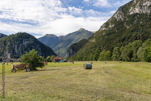 Rural alpine landscape in Valsassina, Lombardy, Italy. Idyllic view of the valley with agricultural fields, hay bales and equipment, surrounded by lush green mountains under a cloudy sky.