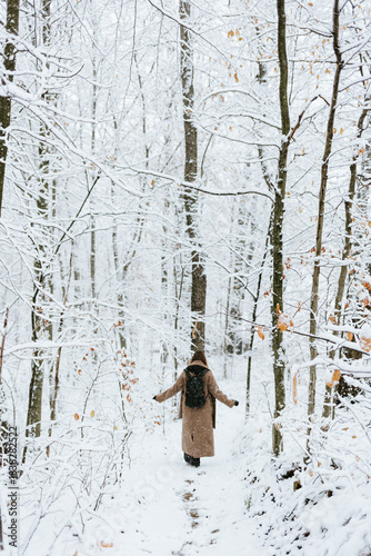 A solitary figure, seen from behind wearing a long, camel-colored coat and a black backpack, walking deliberately along a narrow, snow-covered path cutting through a dense winter forest. 