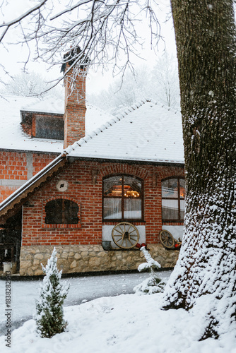 A charming, traditional brick and stone house during heavy snowfall. The focus is on the warm orange-red brick walls and chimney, contrasted sharply with the white, snow-covered tiled roof 