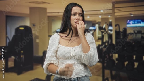 Woman biting fingers in gym near dumbbells and weight rack, hand to mouth nailbiting gesture, wearing white blouse and jeans; anxiety vulnerability.