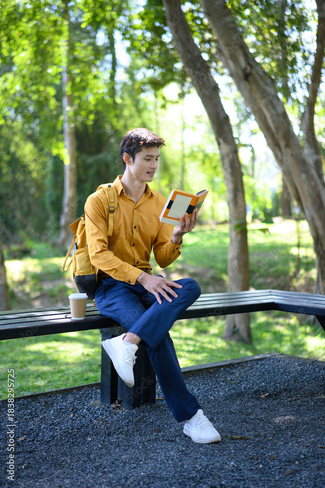 Obraz premium A man sits on a bench in a green park reading a book with a coffee cup