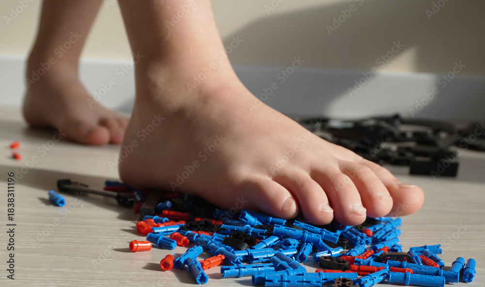 Obraz premium A child's bare foot steps on the scattered parts of a construction kit on the floor. Close-up, parquet, natural light, home scene, emphasis on safety.