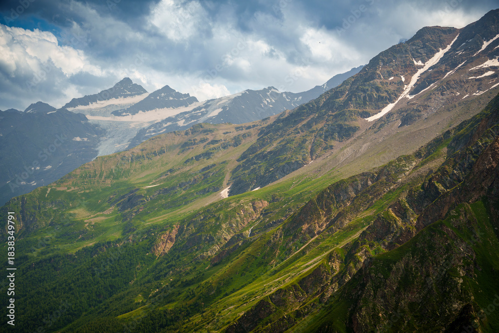 Fototapeta premium Green mountain slope with rocky cliffs and snowy peaks in the distance