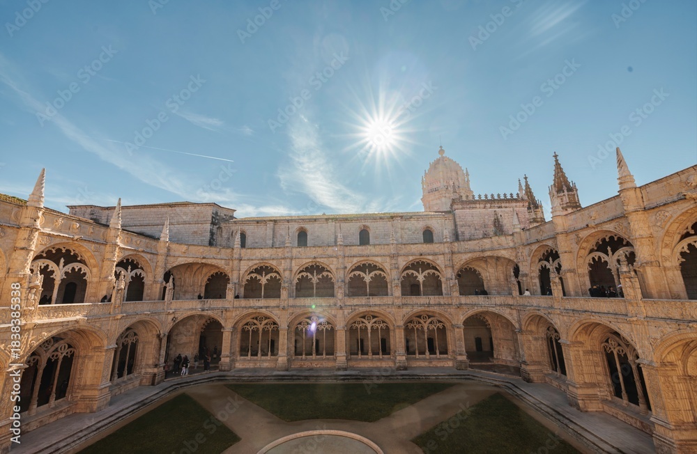 Fototapeta premium Sunlight streams into the courtyard of the Jeronimos Monastery in Lisbon. The ornate architecture of the cloisters gleams under the bright, clear sky during the daytime