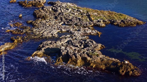 Rugged rocks jut out from the vibrant blue sea, surrounded by lush greenery under a bright, sunny sky. Waves splash against the shoreline, creating a serene coastal scene.