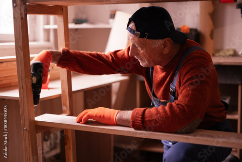 Profession, art and hobby concept. Elderly man carpenter assembling wooden shelf using electric bit drill working on wood in garage. Authentic workshop.