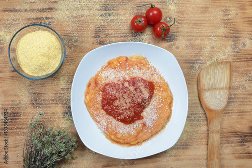 Top view of a plate of traditional Italian polenta with tomato sauce and cheese surrounded by raw ingredients on a wooden table
