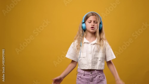 Happy little girl wearing headphones, enjoying music, singing, and dancing with her eyes closed. Child feeling the rhythm on a yellow background