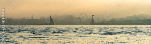 On a foggy winter day in pastel colors, seagulls fly along the waterfront with the port, the Imperia statue, and the Ferris wheel in the background. Konstanz, Baden-Württemberg, Germany.