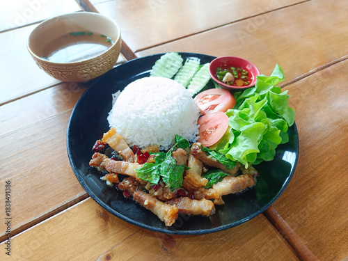  pork with basil topped with rice, vegetables, tomato slices, cucumber slices and dipping sauce in a red bowl, Placed on a black ceramic plate, and soup in a cup, on a wooden table background