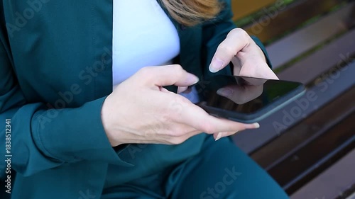 young woman holds smartphone in hands, texting, watching content, sitting on bench in park, close-up view, no face, copy space