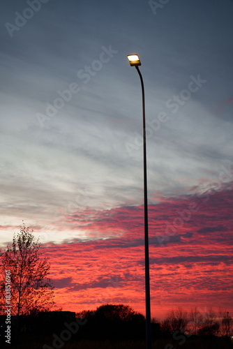 silhouetted streetlamp stands against a vivid red-orange sunset sky, capturing the tranquil transition from day to night with subtle tree outlines in the background, skyline, ambient, peacefulness