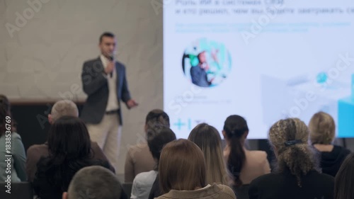 Presenter speaks to audience during conference. Attentive crowd sits facing stage while large screen shows graphics and text. Professional atmosphere emphasizes knowledge sharing collaboration 