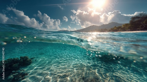 Sunlight reflects on the water surface in the ocean. The underwater scene shows clear blue water and coral formations. Mountains rise in the distance with clouds in the sky.