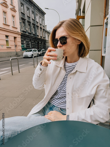 Young blonde woman wearing sunglasses and casual clothes is sitting at table in outdoor cafe, enjoying cup of coffee on cloudy day in city. Woman sipping coffee, wearing sunglasses and casual attire