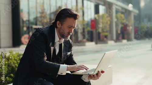 Portrait of elderly corporate CEO man types on laptop keyboard as sits on parapet in downtown. Mature businessman writes email on notebook computer working outside office building
