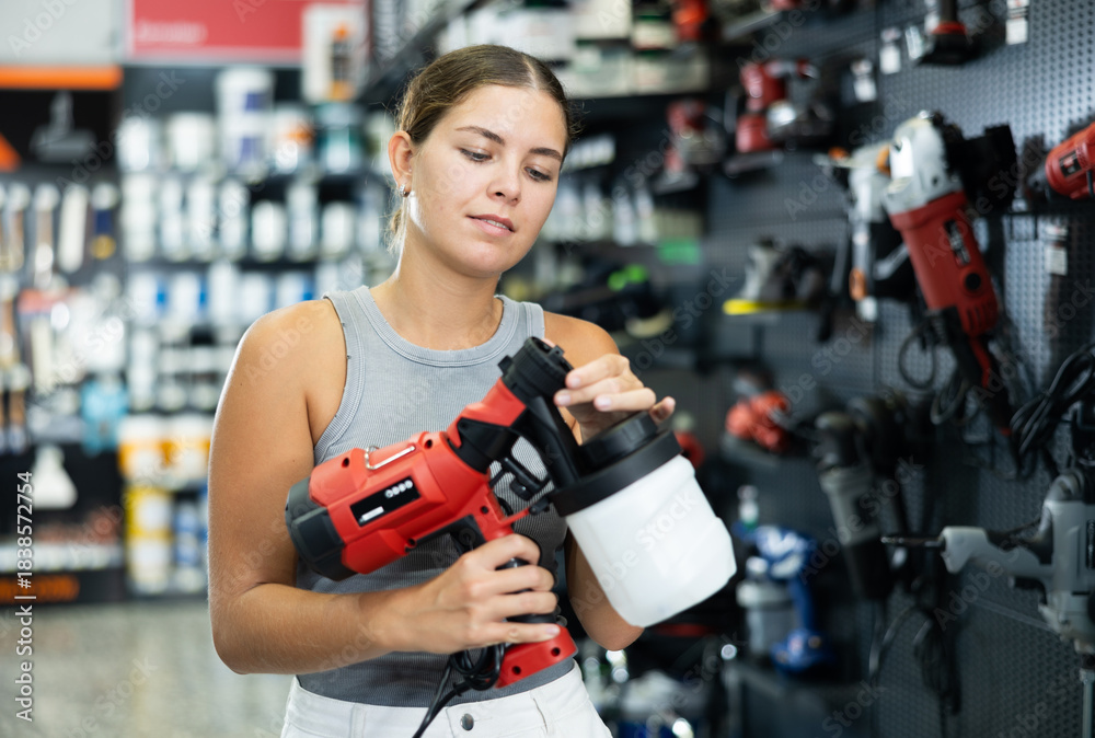 Fototapeta premium Beautiful young girl planning home renovation and choosing paint sprayer in hardware store