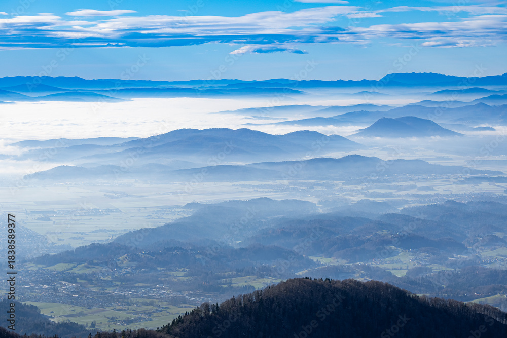 Obraz premium Wide panoramic view across valleys filled with dense fog, showcasing layered mountain ridges and the hill town of Kamnik in the foreground