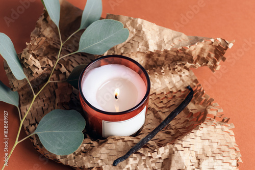Lit candle in red glass with eucalyptus leaves and textured paper on warm orange background. Top view