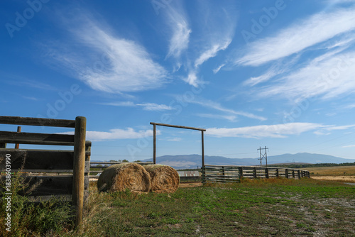 Country ranch setting with old wooden corral fence and metal gate with round straw or hay bales and loading chute for livestock under big blue sky with clouds