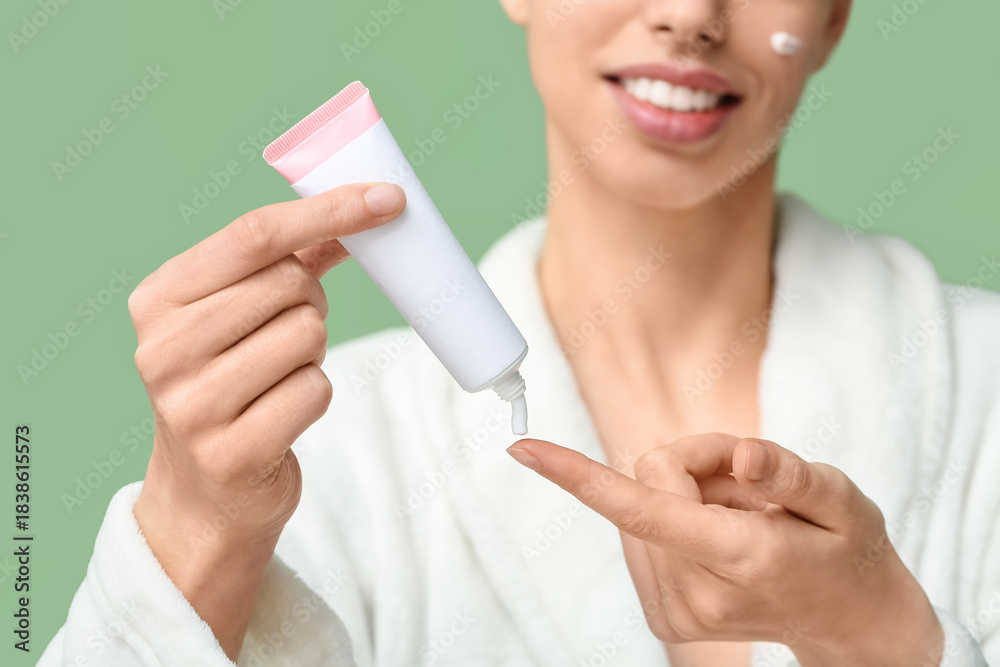 Naklejka premium Beautiful young woman in robe with tube of face cream on green background
